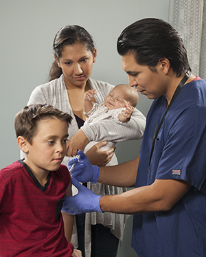 Health care provider giving boy an injection, and in background mother holding baby.